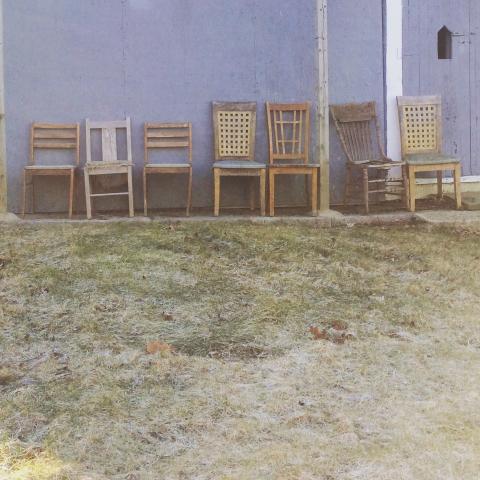 A variety of aged wooden chairs lined up outside against a house.